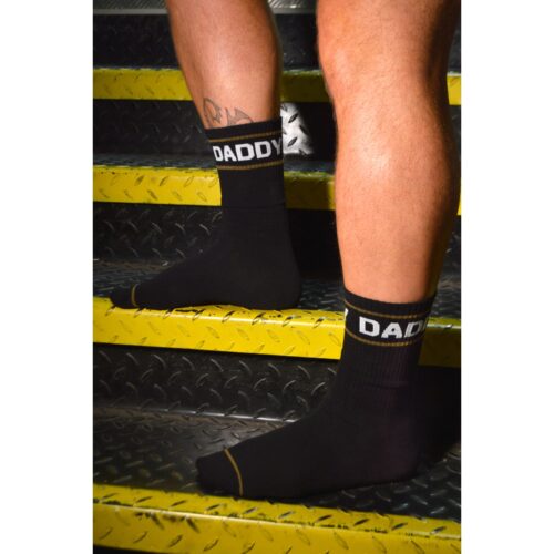 Black socks with 'DADDY' on the cuffs, worn on a person’s feet on yellow stairs