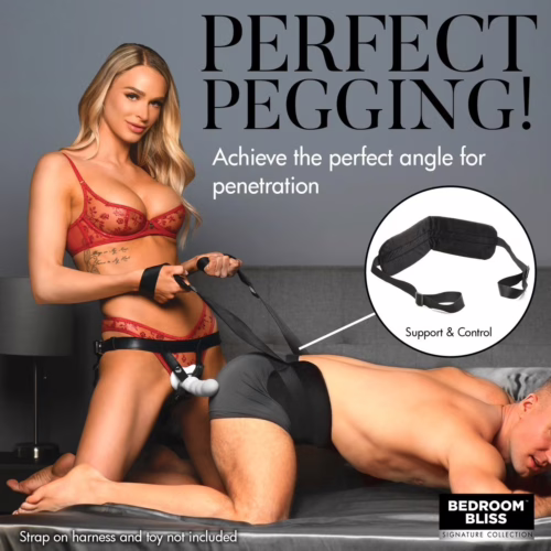 A woman in red lingerie holds a harness for pegging, with a man in shorts on a couch