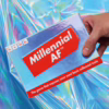 Discover your millennial traits with the fun 'Millennial AF' card game, now available at The Loving Company. Hand holding 'Millennial AF' card game, against a holographic background